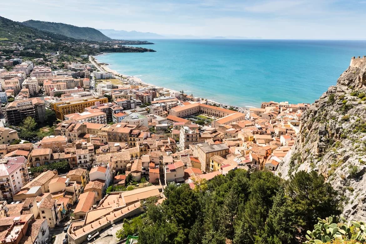 A high-angle view captures the city of Cefalù, with its dense red-tiled roofs and the sea stretching to the horizon.