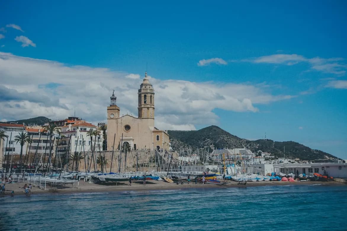 The historic church of Sant Bartomeu and Santa Tecla stands at the edge of a sandy beach, with a few sailboats and yachts moored nearby.