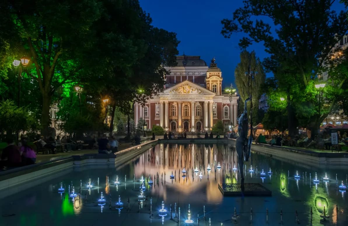 The Ivan Vazov National Theatre is beautifully illuminated at night and reflected on a large fountain in a city park.