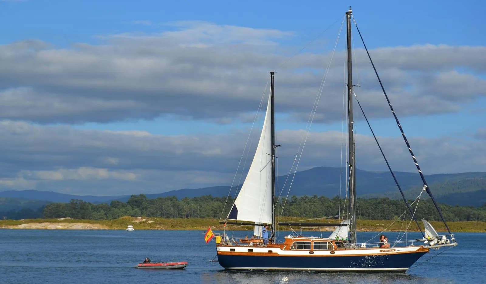 A large sailboat with a white sail cruises on the calm blue water, with a lush green hillside in the background.