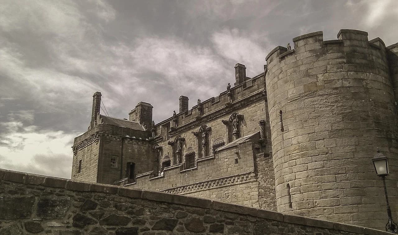 A large, ornate stone building with fortified walls and crenelations is viewed from a low angle, showcasing its medieval architecture.
