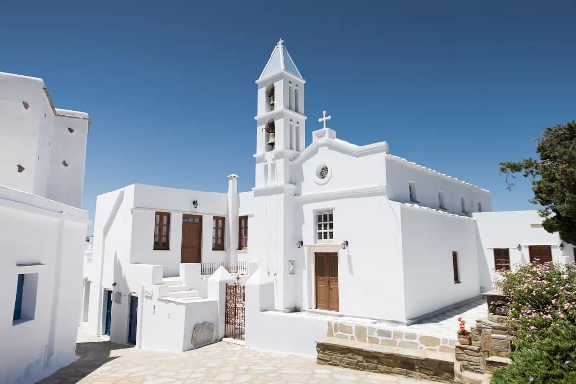 A narrow, white-washed alleyway in Tinos is lined with traditional Cycladic buildings, with a few small cafes and shops.