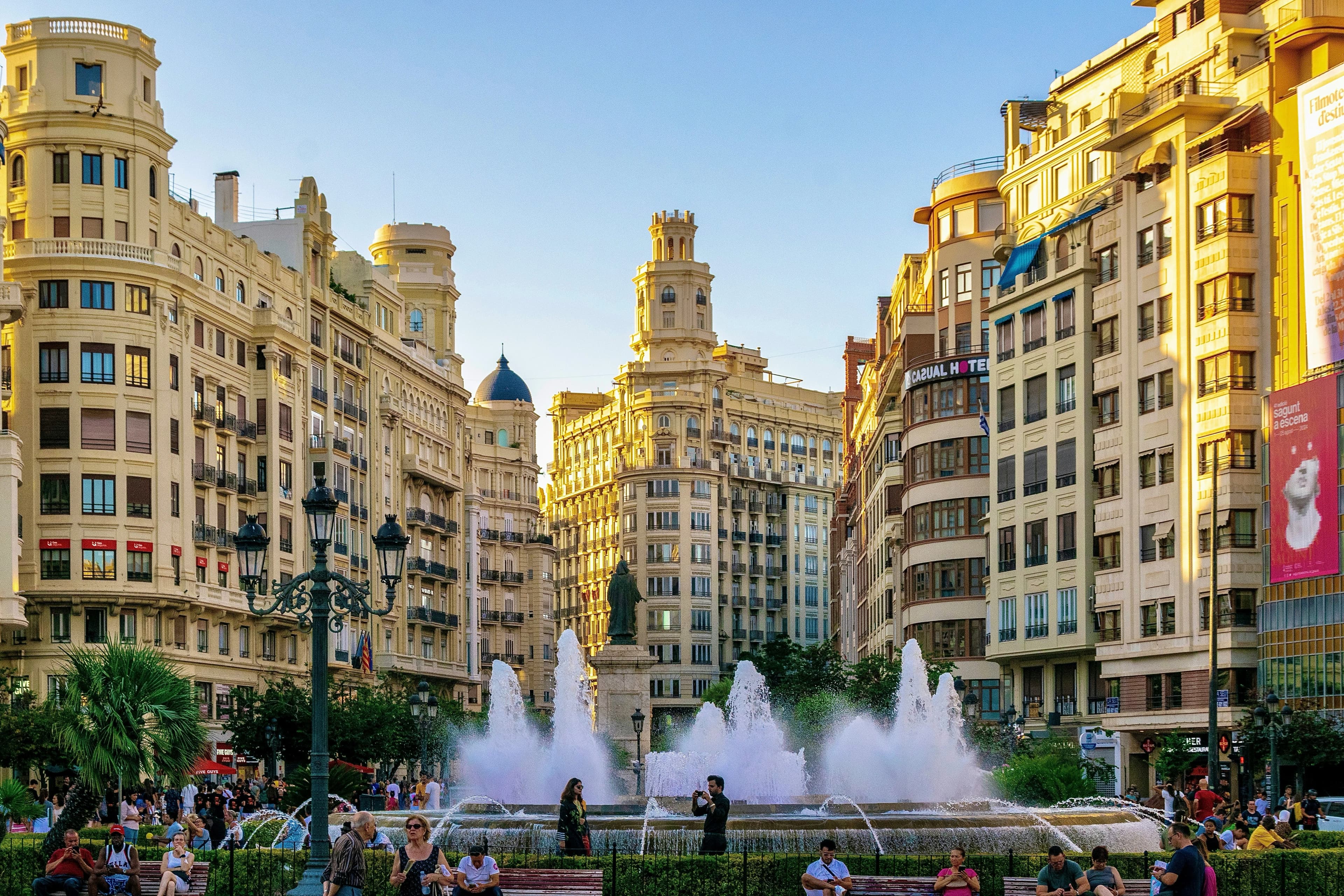 People gather in the Plaza del Ayuntamiento, Valencia's main square, with a large fountain and elegant, neoclassical buildings.