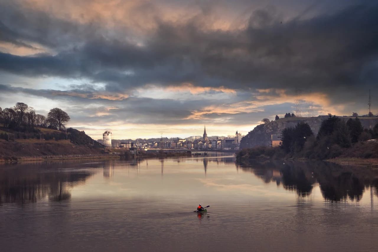A person in a kayak on a calm river with a scenic view of Waterford in the background at sunset.