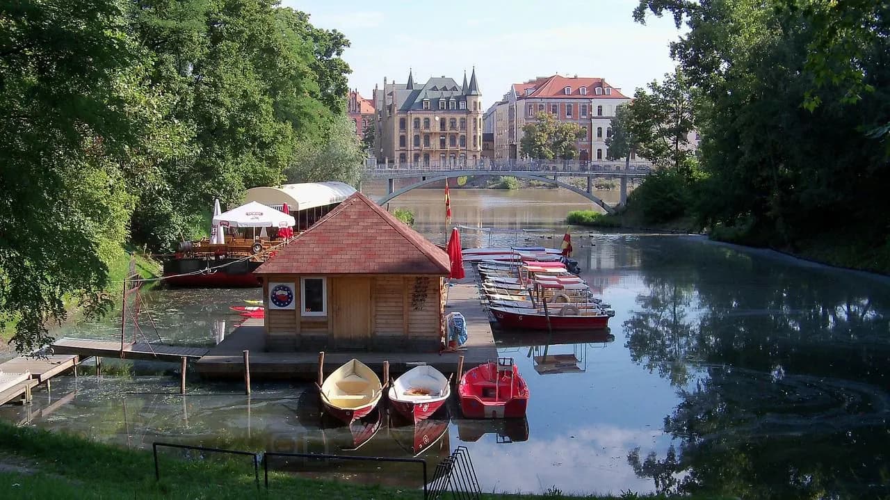 A quaint wooden boat house with colorful boats moored nearby is nestled on a tranquil river, with a historic bridge in the background.